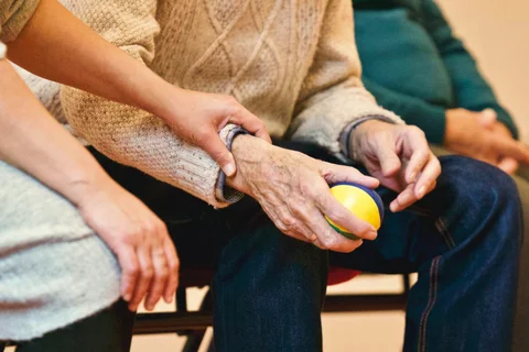 An older couple are seen sitting together. One is holding the other's wrist reassuringly.