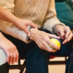 An older couple are seen sitting together. One is holding the other's wrist reassuringly.